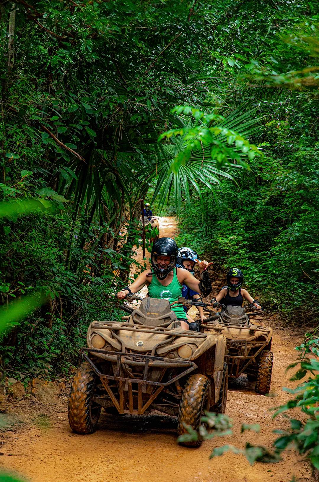 Un grupo de personas en cuatrimotos en un sendero de tierra, atravesando un denso bosque verde, con cascos y emocionados.