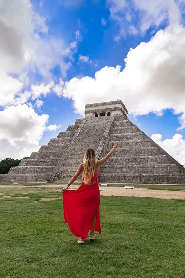 Woman in a red dress walking towards the Chichen Itza pyramid under a vibrant blue sky with scattered clouds.