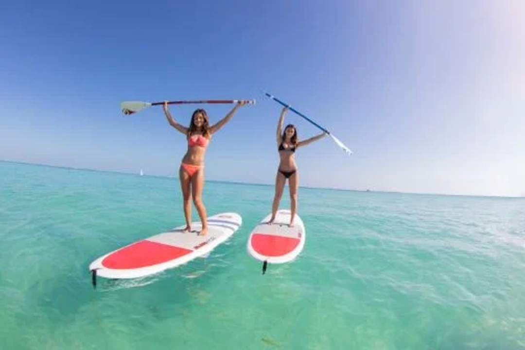 Two women in bikinis stand on paddleboards, holding paddles above their heads in clear blue water.