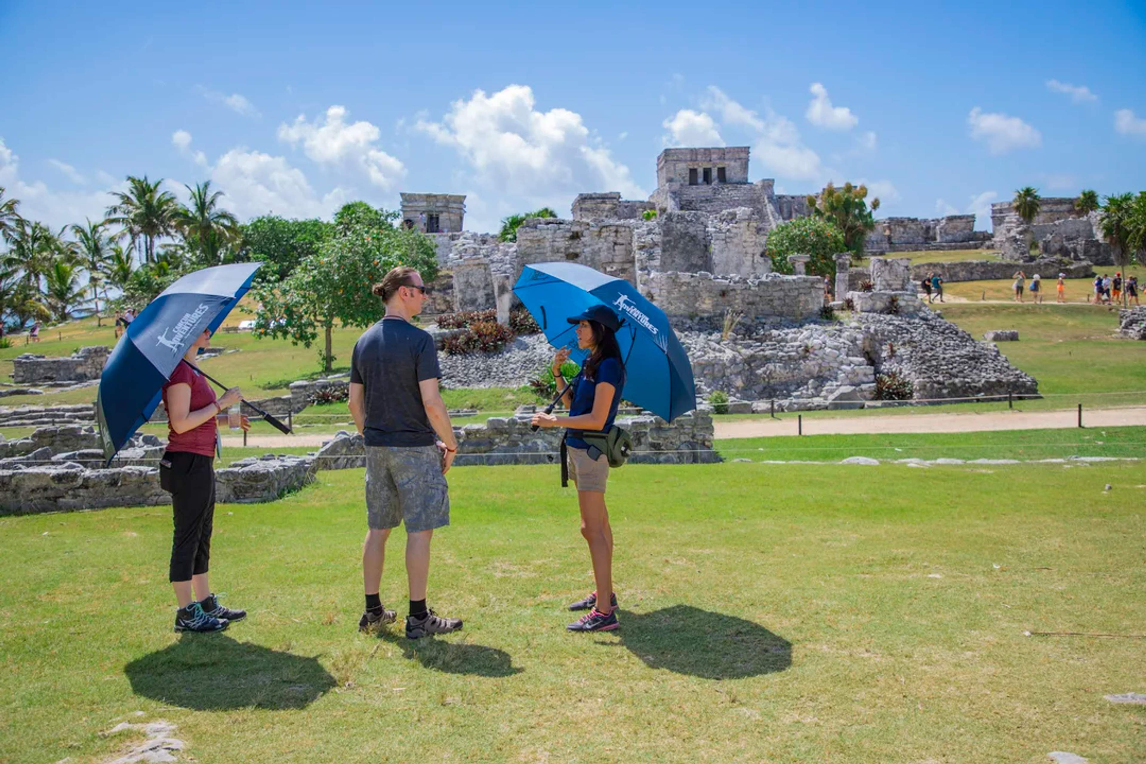 Turistas recorren antiguas ruinas mayas en Tulum bajo el sol con guía y sombrillas protectoras.