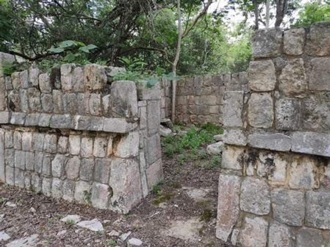 A photograph of the Casa de las Cabacitas at Chichen Itza, featuring partially preserved stone walls of an ancient structure. The site is surrounded by greenery and trees. Photo credit: Giovanni Agostino Frassetto.