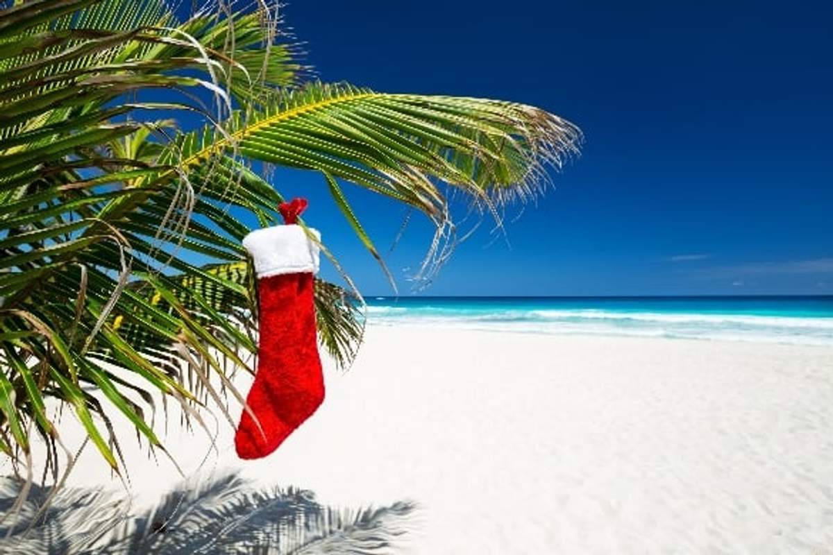 Calcetín navideño rojo colgando de una palmera en una playa de arena blanca, con agua turquesa y un cielo azul claro de fondo.