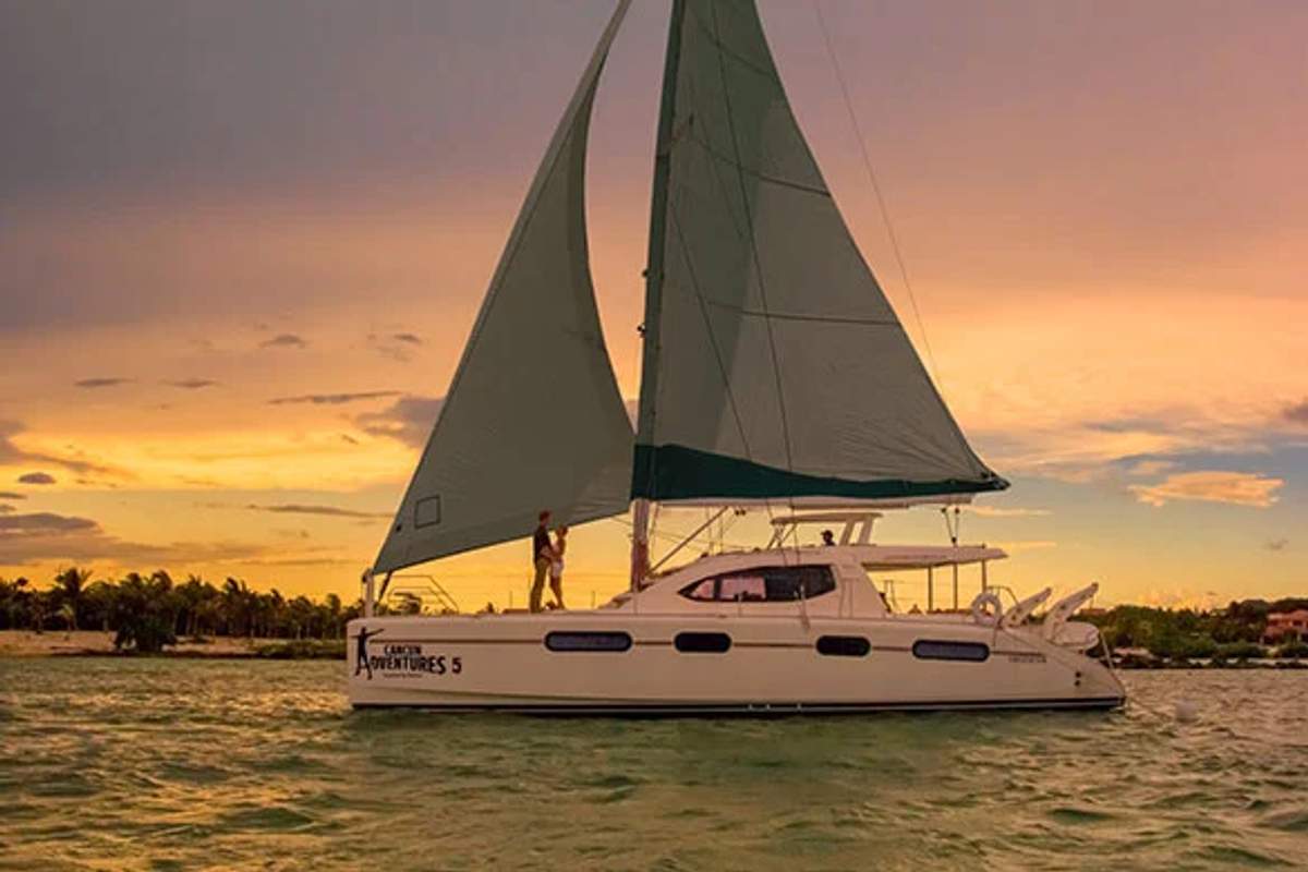 A sailboat cruising at sunset in Cozumel, with a person standing on deck and the sky painted in warm hues.