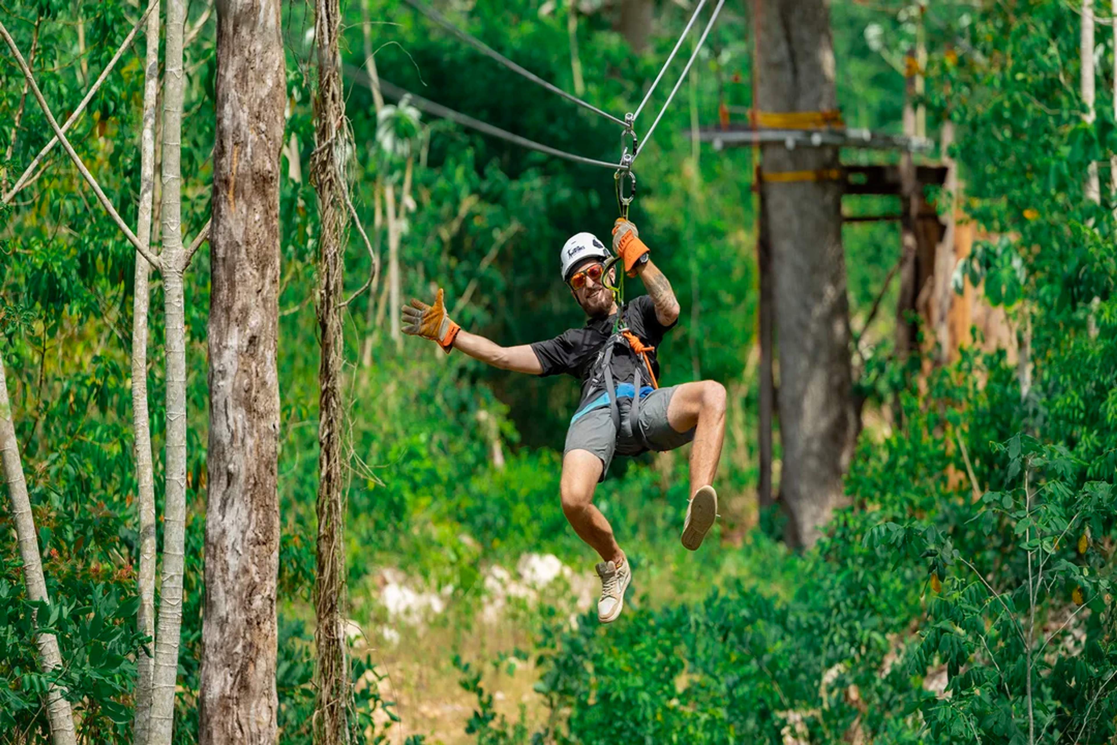 Hombre disfruta de una emocionante tirolesa en la selva, sonriendo y saludando en el aire.