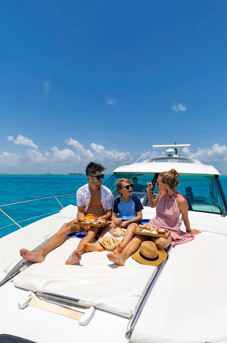 Una familia disfrutando de aperitivos en la cubierta de un yate bajo un cielo azul claro, con agua turquesa de fondo.