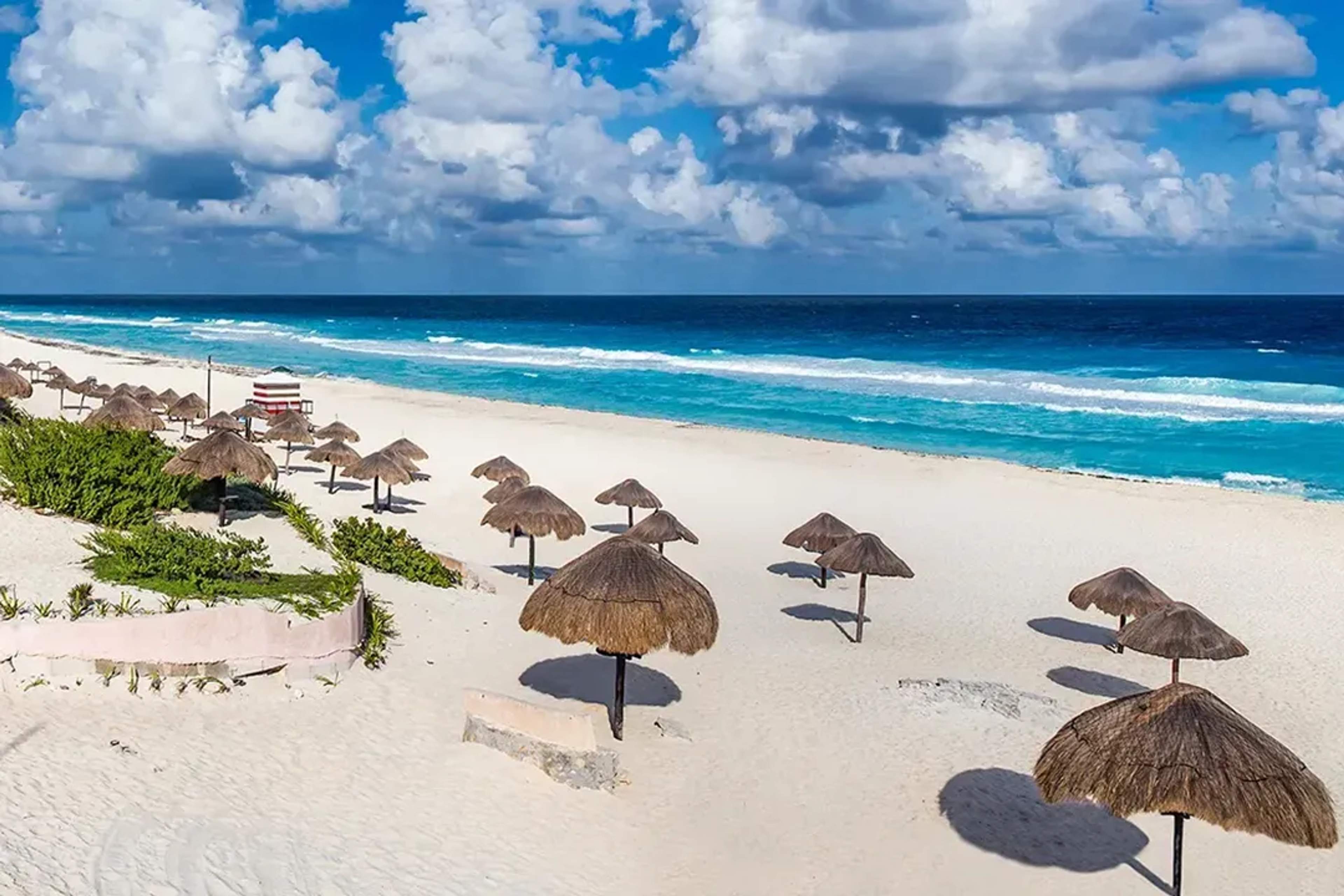 Playa de Cancún con arena blanca, palapas y olas turquesa bajo un cielo brillante con nubes dispersas