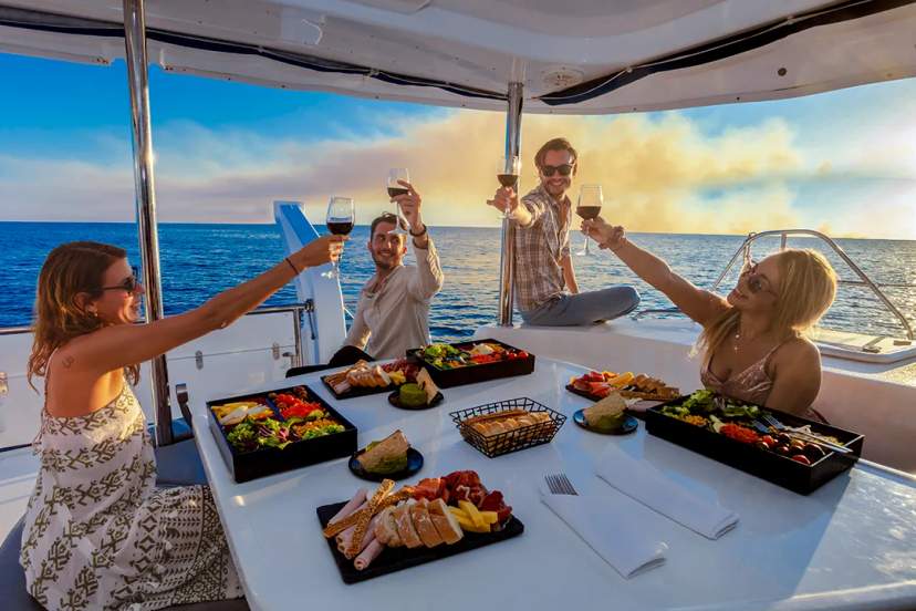 Amigos disfrutan de una comida gourmet y bebidas en un paseo en barco en Cancún, con el océano y el atardecer de fondo.