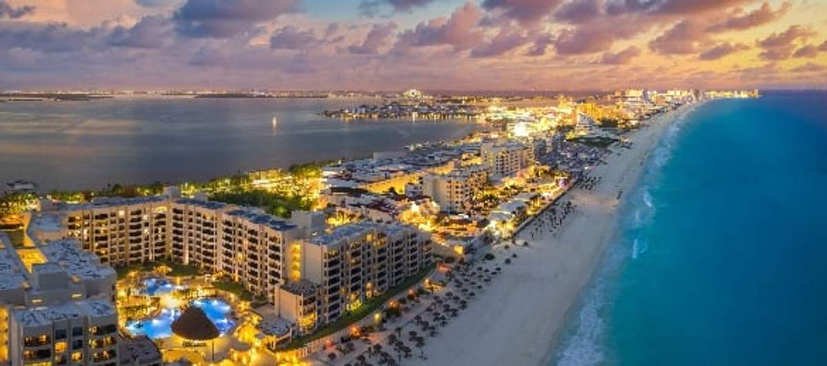 Vista aérea de una ciudad costera al atardecer, con numerosos hoteles iluminados que bordean una playa de arena junto a aguas turquesas a la derecha y una bahía tranquila a la izquierda, bajo un cielo con nubes dispersas.