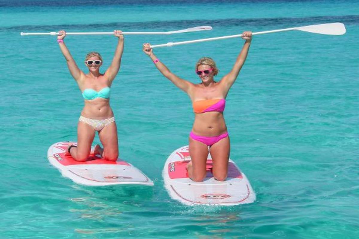 Dos mujeres en bikini disfrutando de un día de paddle surf en aguas cristalinas y turquesas. Ambas están de pie sobre sus tablas de paddle, levantando sus remos con entusiasmo. La escena transmite diversión y energía en un entorno tropical.