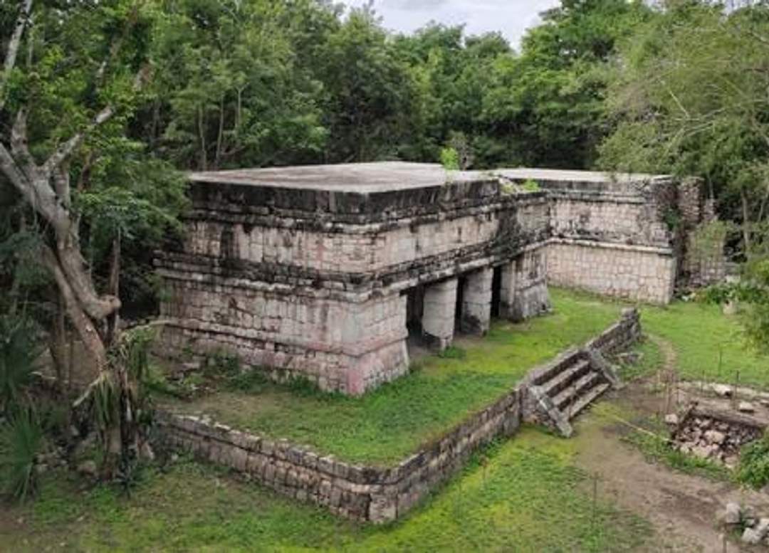 A photograph of the Temple of the Owls at Chichen Itza, featuring an ancient stone structure with multiple columns and a staircase leading to the entrance. The temple is surrounded by lush greenery. Photo credit: Giovanni Agostino Frassetto.