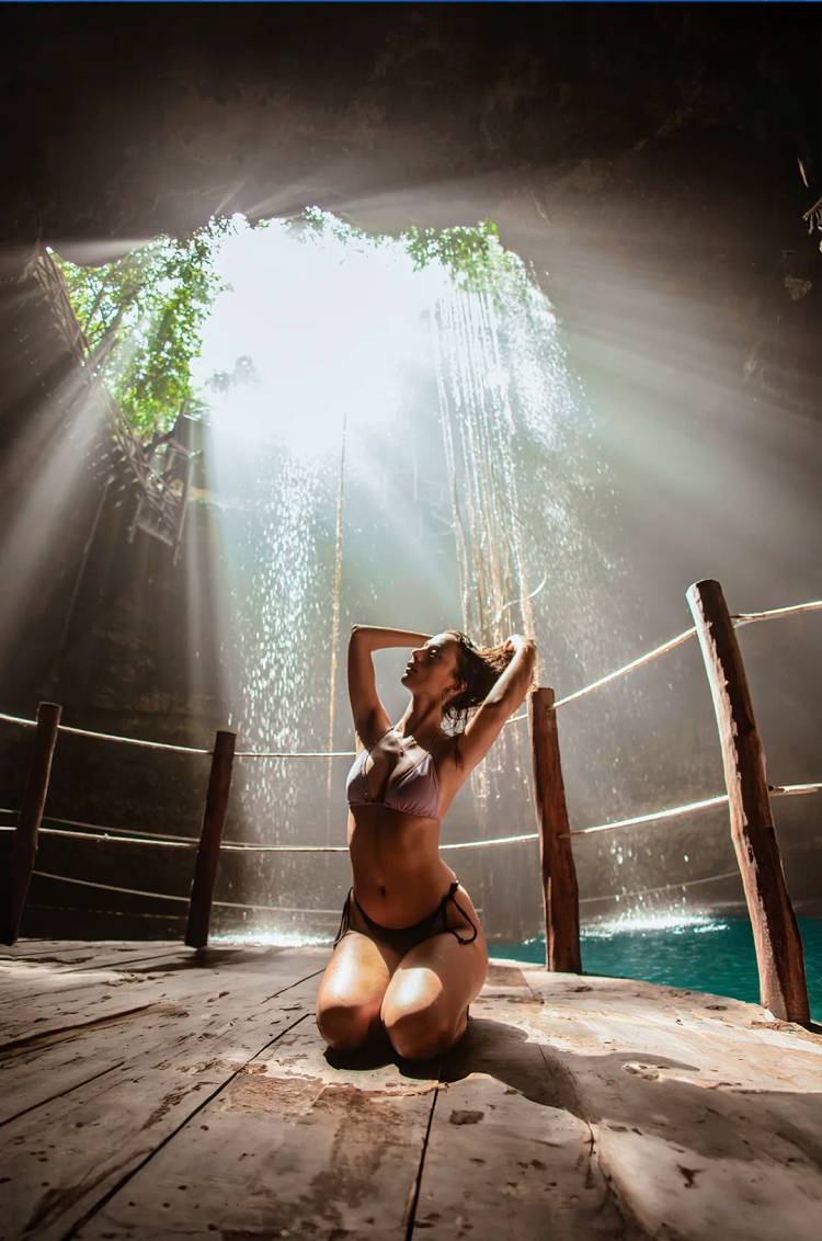  A woman kneels in a cenote, bathed in sunlight streaming through the cave's opening, creating a magical, serene atmosphere.