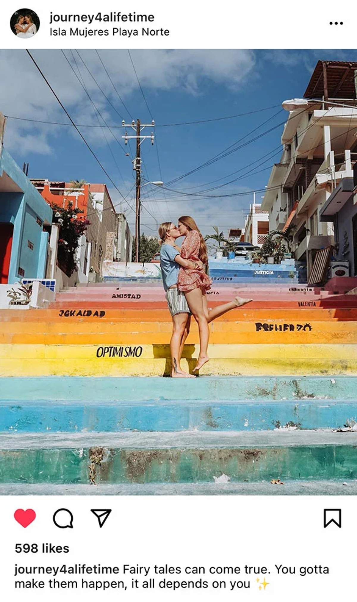 Una pareja besándose en unos escalones de arcoíris coloridos en Isla Mujeres, con palabras como "AMISTAD," "OPTIMISMO," y "VALENTIA" escritas en los escalones.