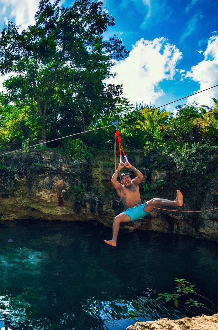 A man in blue shorts ziplining over a clear water cenote, smiling and preparing to drop into the water.