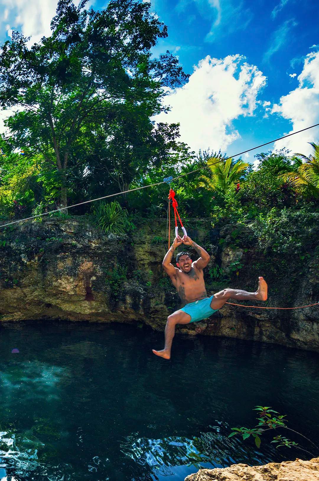 Un hombre con shorts azules haciendo tirolesa sobre un cenote de agua clara, sonriendo y preparándose para caer al agua.