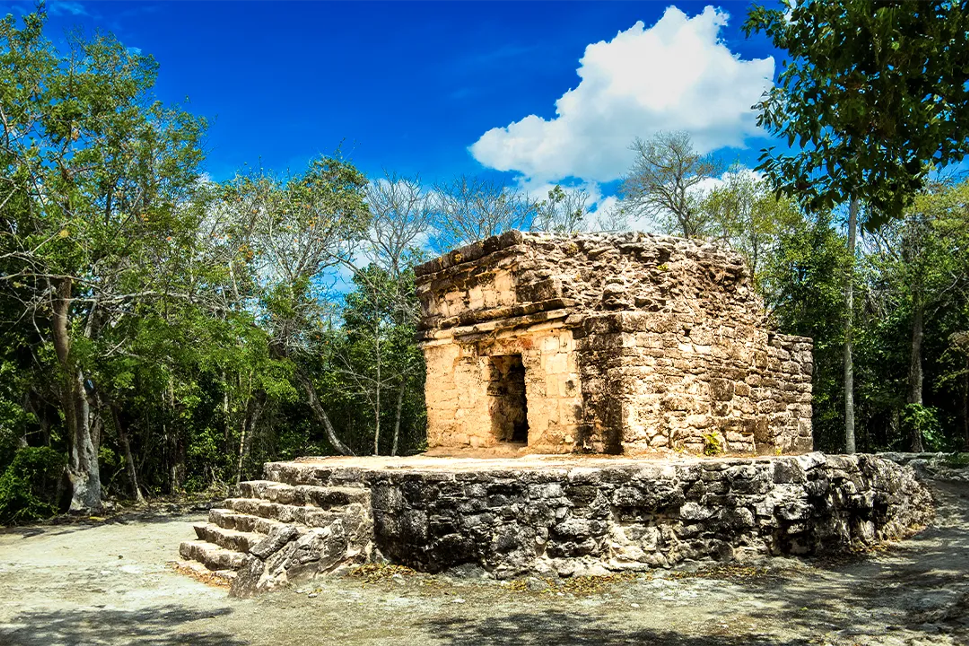 Ancient stone structure of Old Chichen surrounded by lush jungle under a bright blue sky.