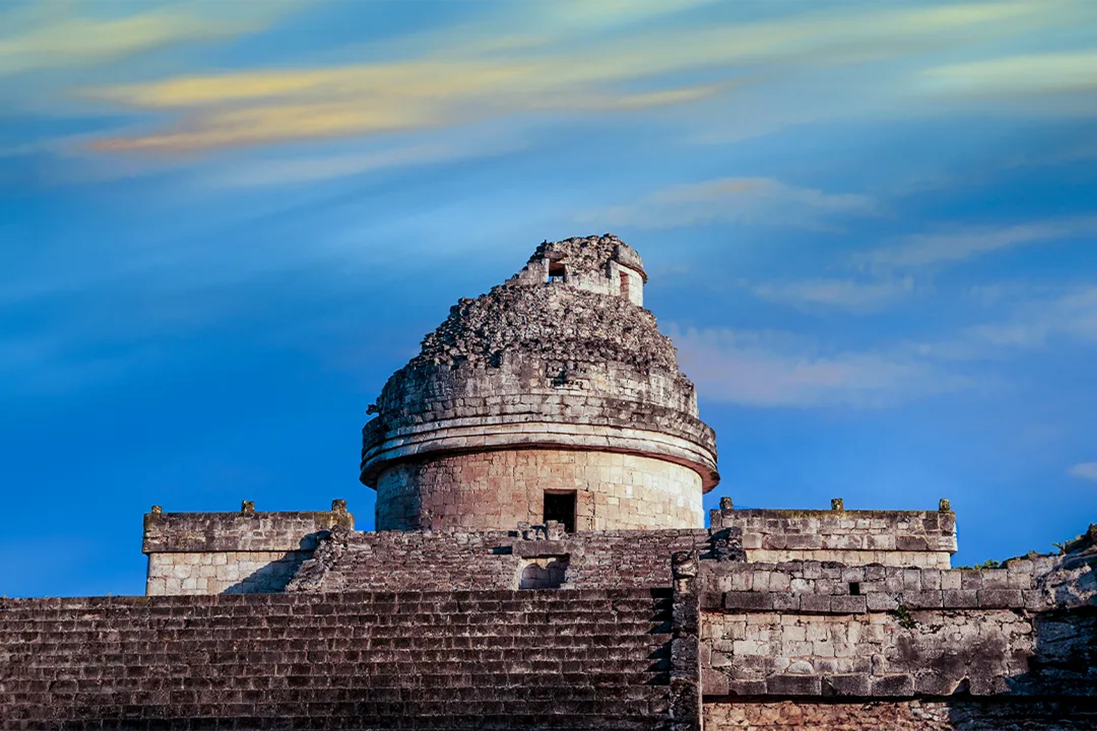 The observatory at Chichen Itza, known as El Caracol, under a colorful sky.