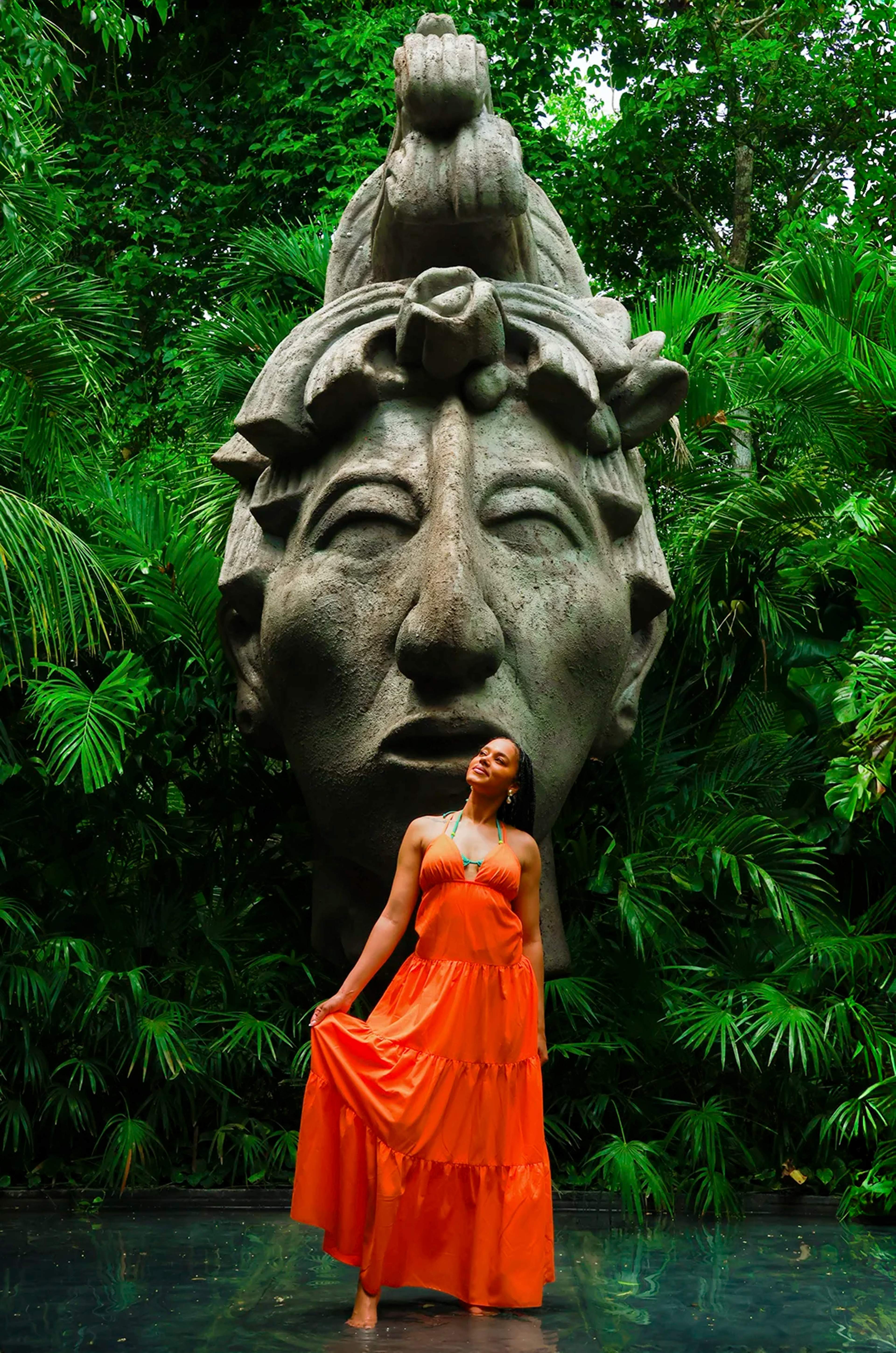 Mujer con vestido naranja posa frente a cabeza maya gigante, rodeada de verde selva frondosa.
