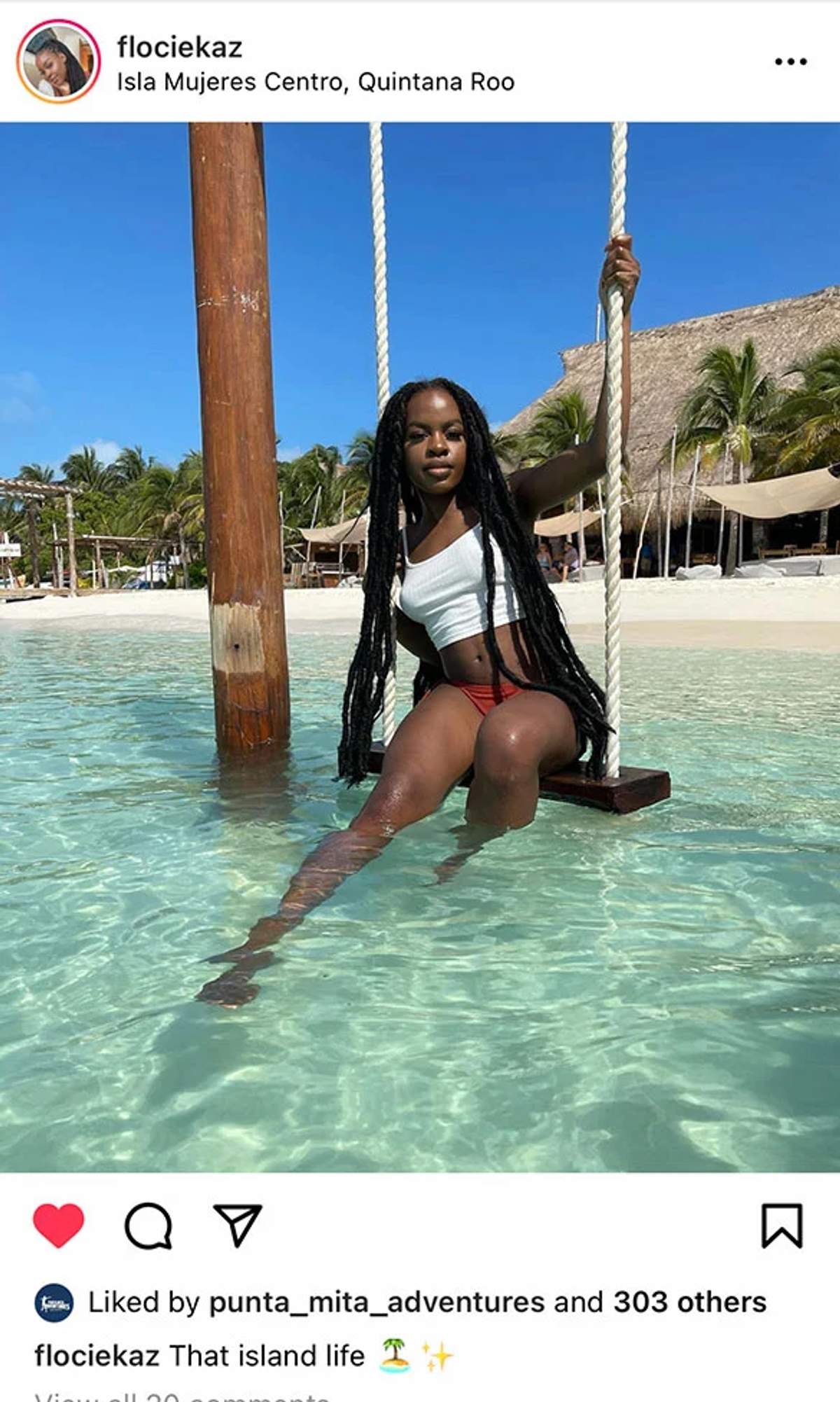 Una mujer con un top blanco y un bikini rojo se sienta en un columpio sobre agua clara y turquesa con una playa tropical y palmeras en el fondo en Isla Mujeres Centro, Quintana Roo. 