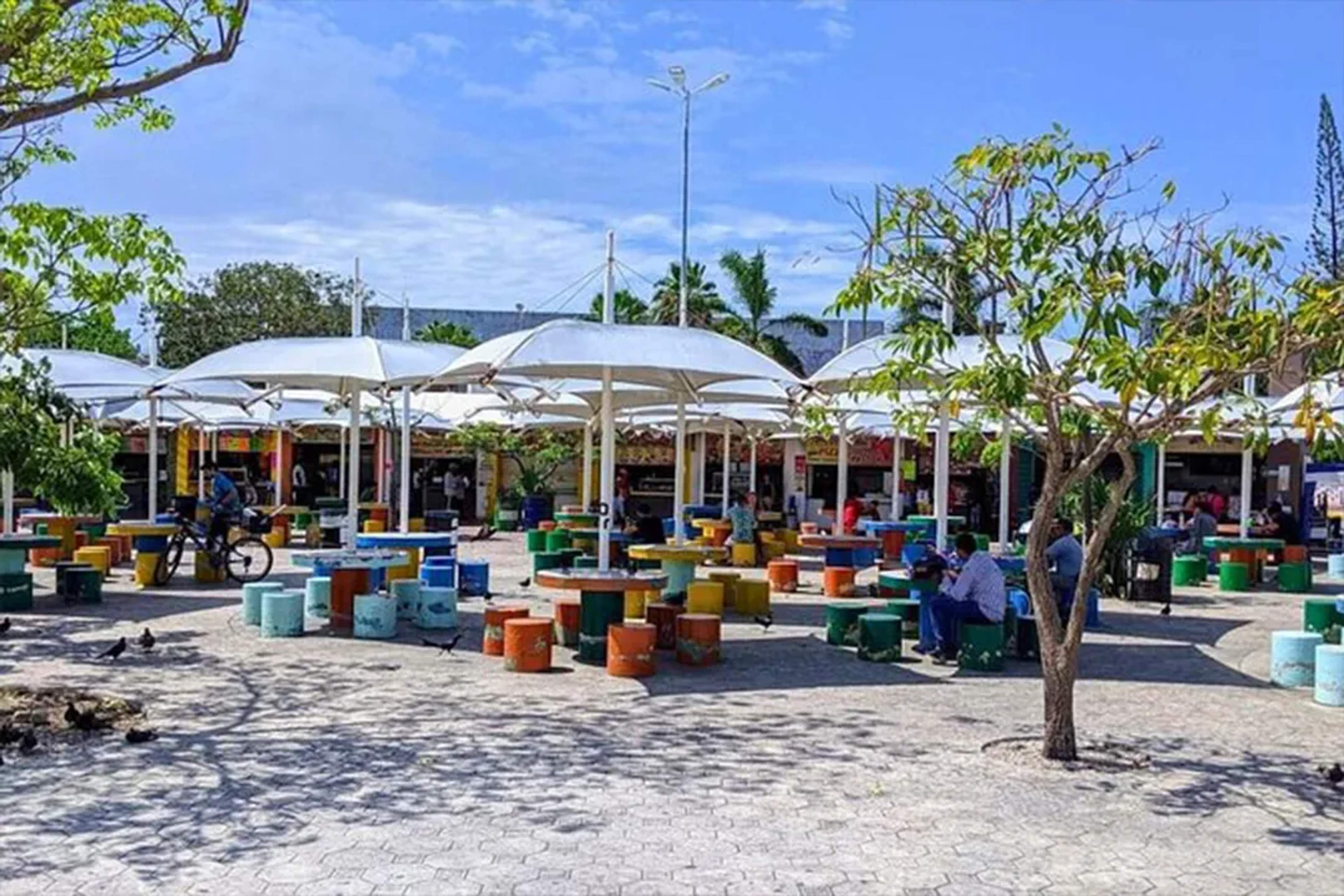 Colorida plaza de comidas al aire libre con mesas redondas, sombrillas y algunas personas.