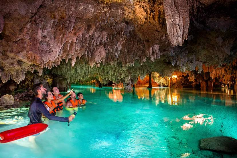 Group swimming in illuminated underground cenote cave with turquoise water and stalactites during Riviera Maya tour