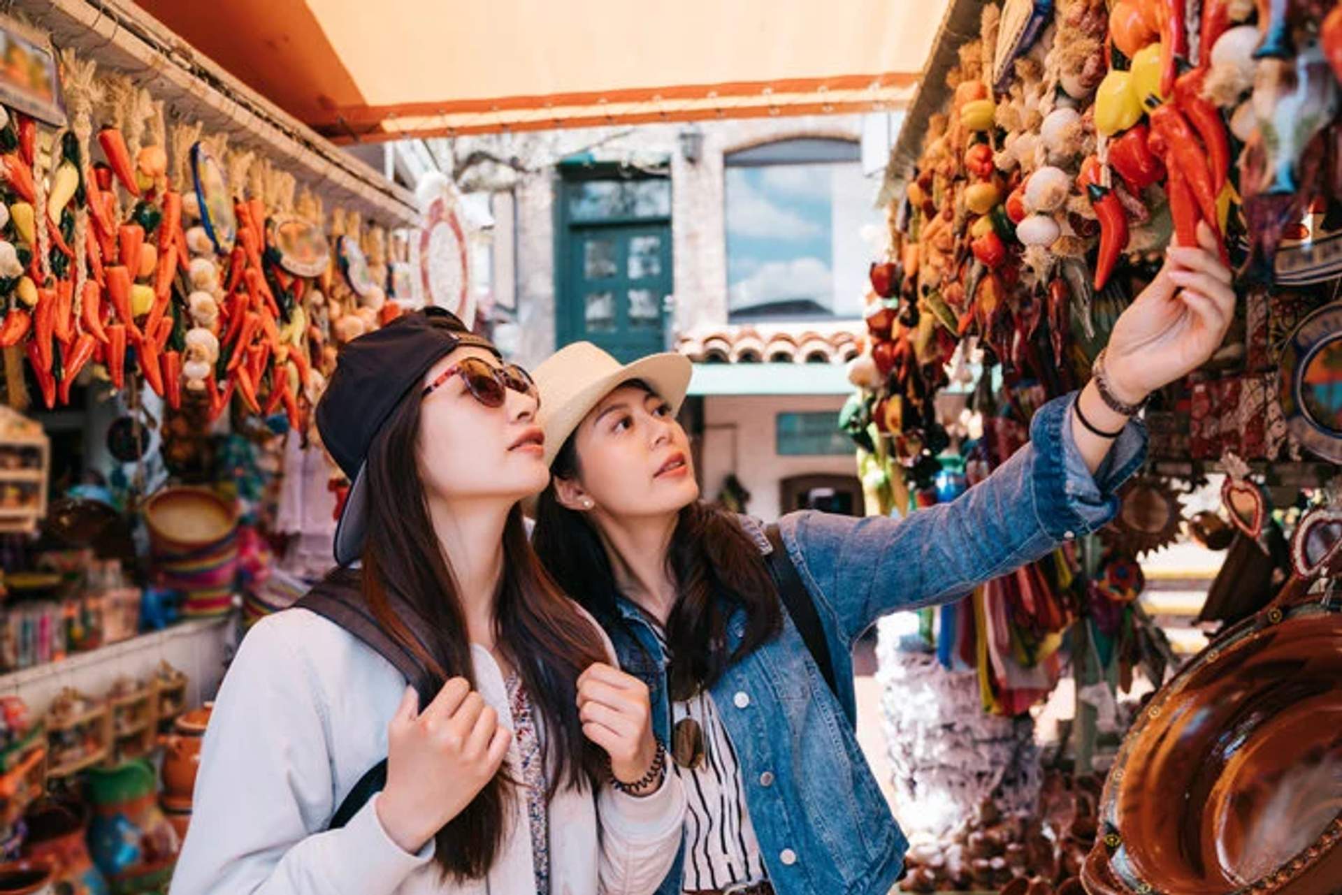 Two women in hats and sunglasses explore a colorful Mexican market, examining hanging decorations and souvenirs.