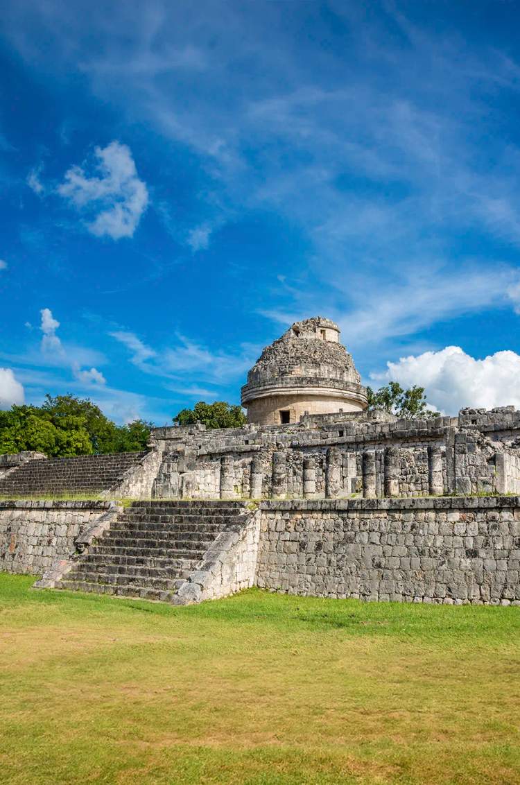 The ancient Mayan observatory "El Caracol" at Chichen Itza, under a bright blue sky with green grass in the foreground.
