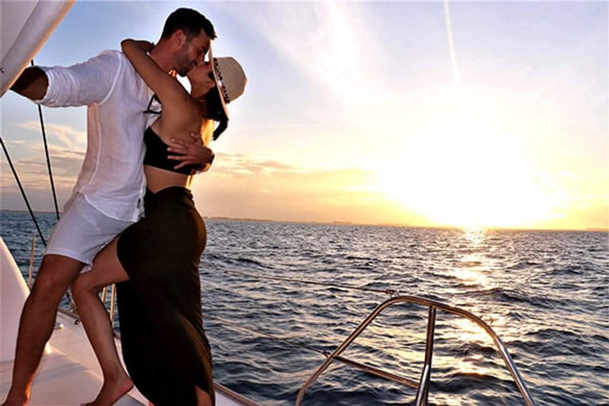 A couple shares a kiss on a luxury boat during a sunset cruise in Cancun, with the sun setting over the ocean in the background.