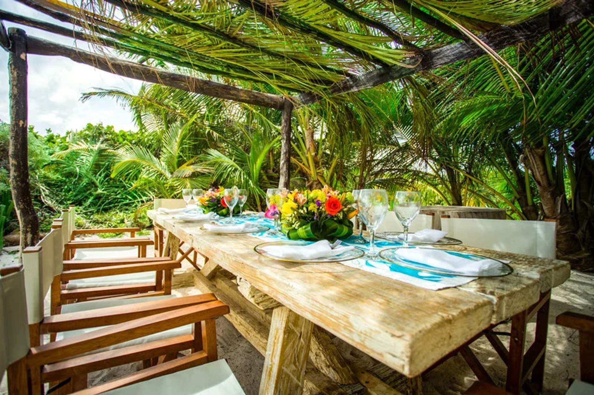 Mesa rústica al aire libre con sillas de madera, adornada con flores coloridas y copas, rodeada de palmeras tropicales.