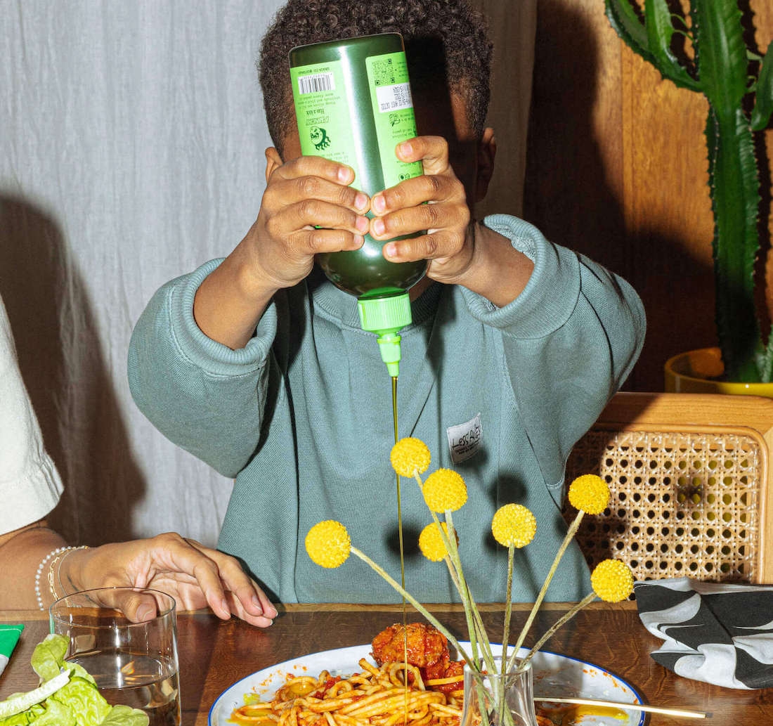 Kid using Graza squeeze bottle at kitchen table with pasta 