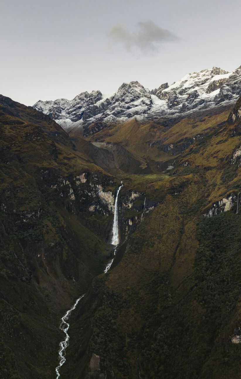 Una cascada alta cae por la ladera verde y empinada de un valle hacia un río sinuoso, bajo cumbres nevadas de la cordillera Vilcabamba, en el lado del Salkantay del valle de Ahobamba, Perú