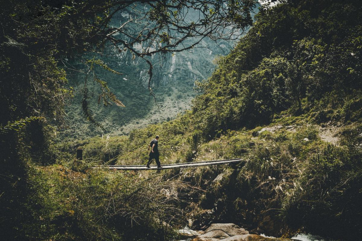 Cliente de AAV cruzando un puente colgante angosto sobre el río Ahobamba, rodeado de bosque nuboso bajo las laderas empinadas del valle, trek Ahobamba a Machu Picchu, Perú