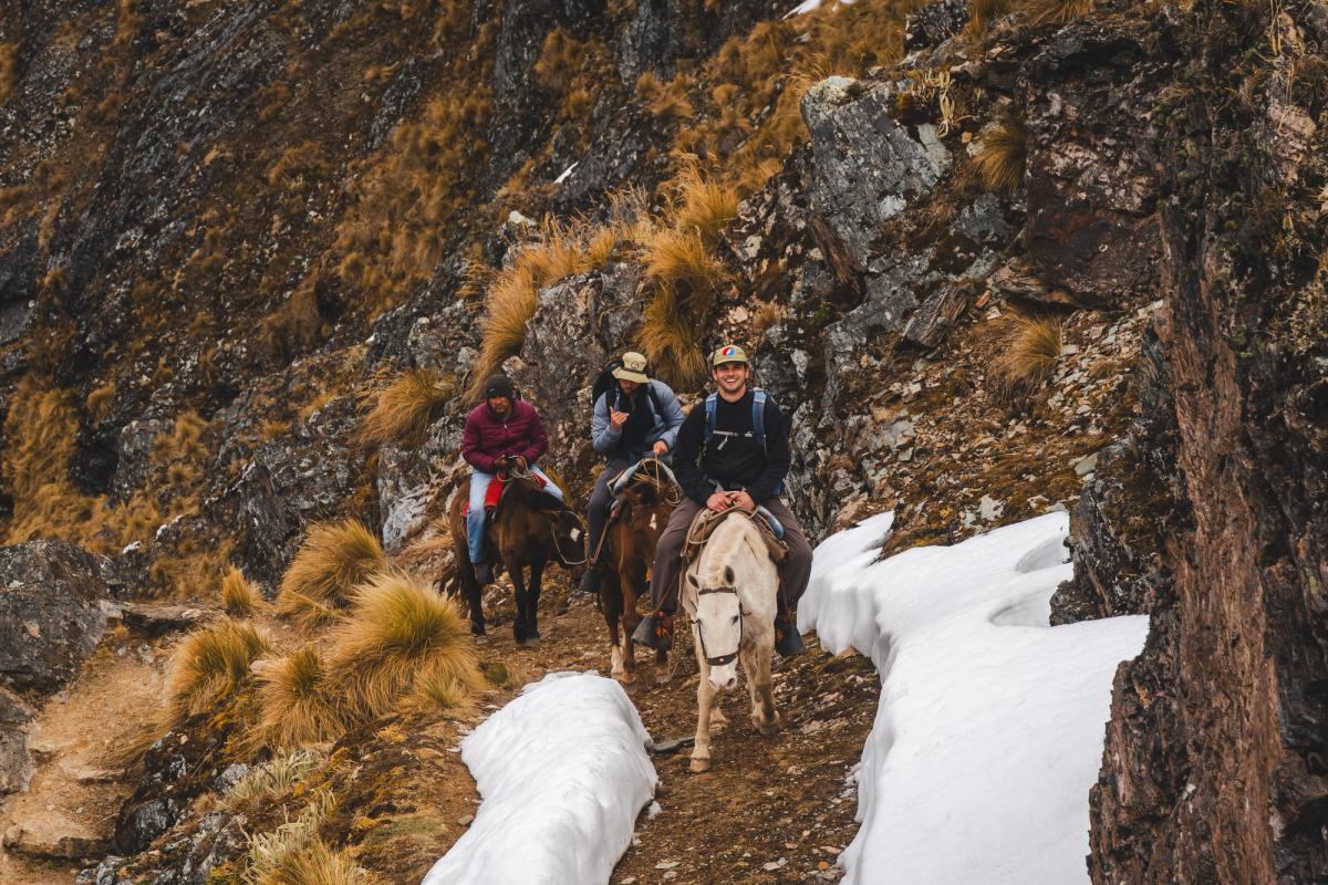 AAV clients riding horses up a narrow rocky trail between patches of snow and golden grass on the climb to Palkay Pass, Ahobamba trek, Peru