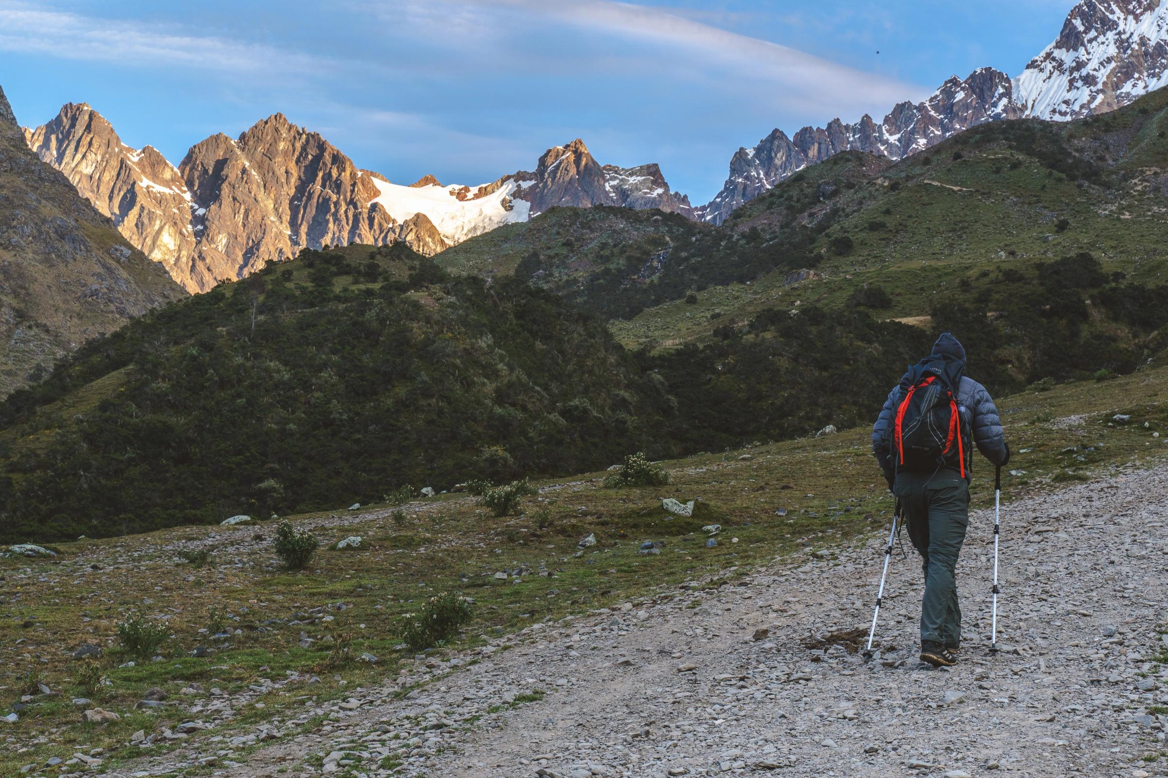Excursionista con bastones de trekking caminando por un sendero rocoso andino hacia montañas nevadas iluminadas por la luz temprana de la mañana.