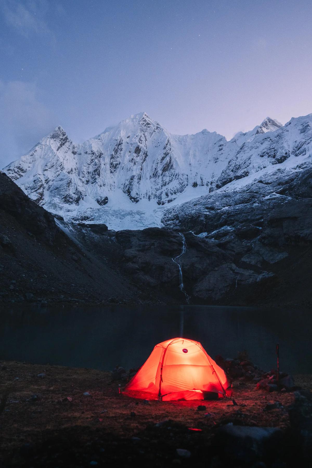 Una carpa naranja brilla al atardecer junto a una laguna, bajo una cascada y picos nevados, con un cielo azul profundo en la Cordillera Huayhuash, Perú