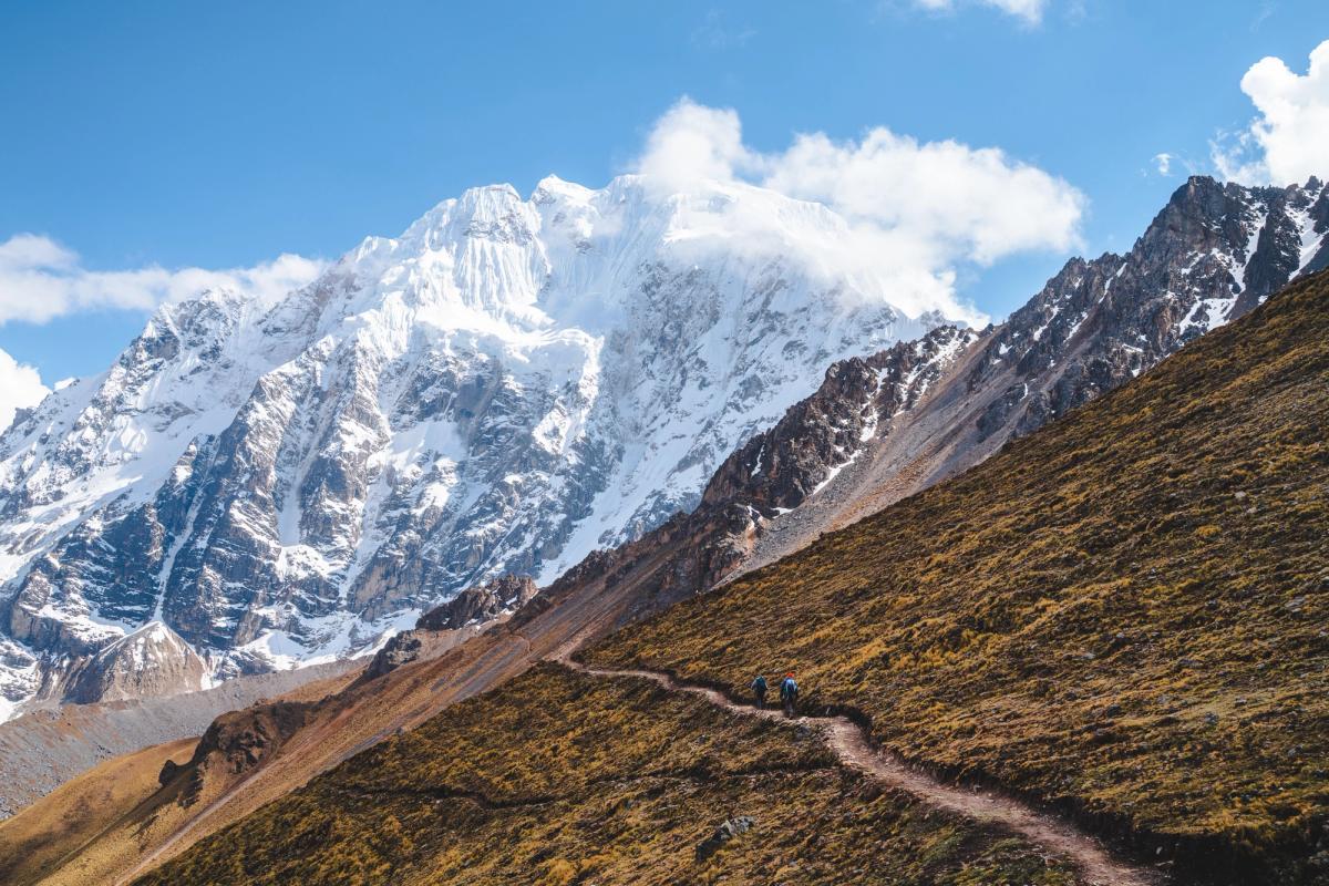 Dos caminantes de AAV en un sendero angosto que cruza una ladera de pajonal dorado, uno de ellos saludando, bajo la enorme cara glaciar del Nevado Salkantay y un cielo azul, Día 1 del trek Ahobamba, Perú