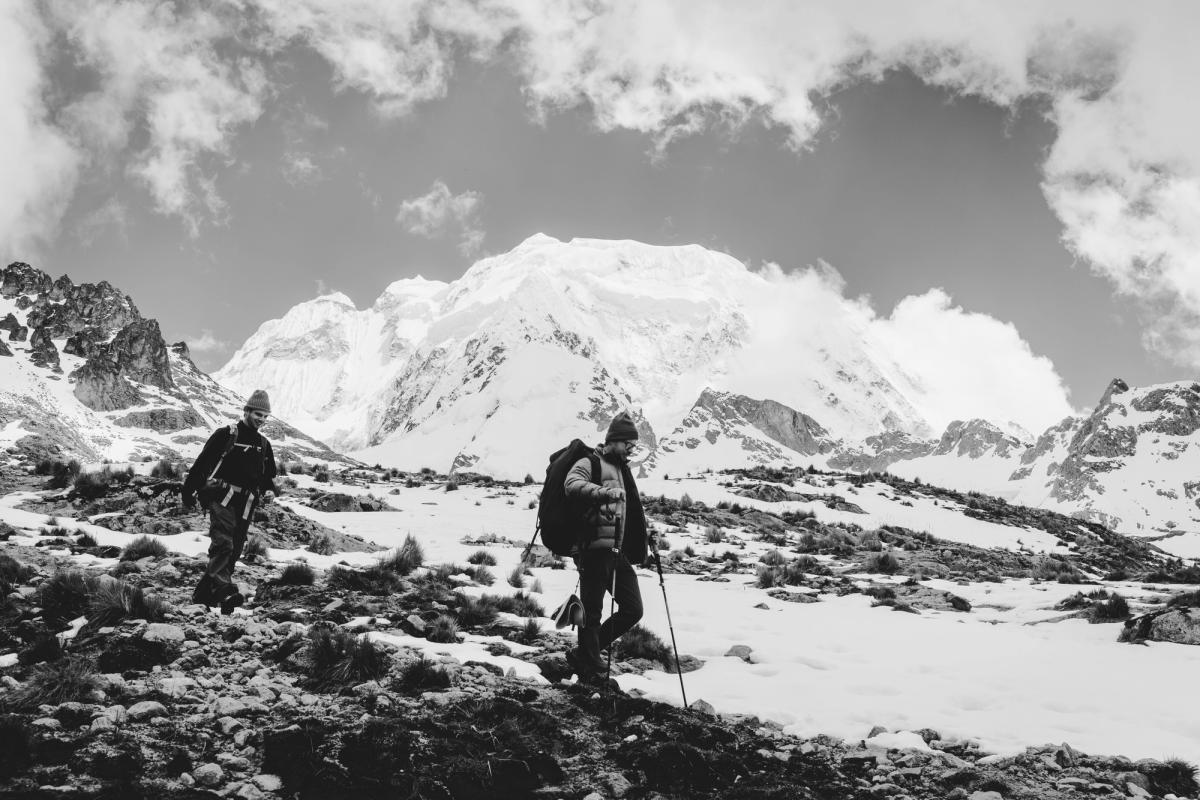 Foto en blanco y negro de dos caminantes de AAV bajando del Abra Incachiriaska sobre nieve y pajonal rocoso, con la cara glaciar del Nevado Salkantay detrás, Día 1 del trek Ahobamba, Perú