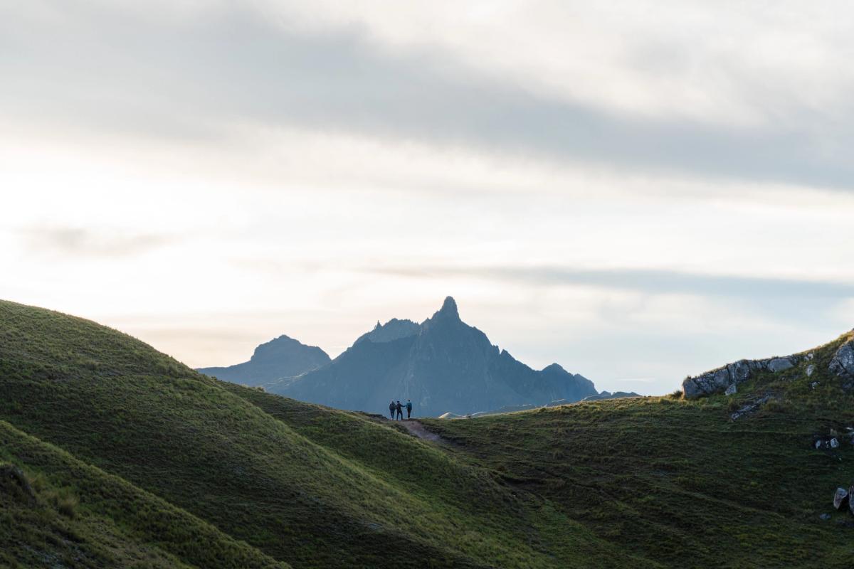 Hikers walking along a high Andean ridge during the Ancascoca Expedition, with a dramatic jagged mountain peak rising in the background under soft morning light in April.