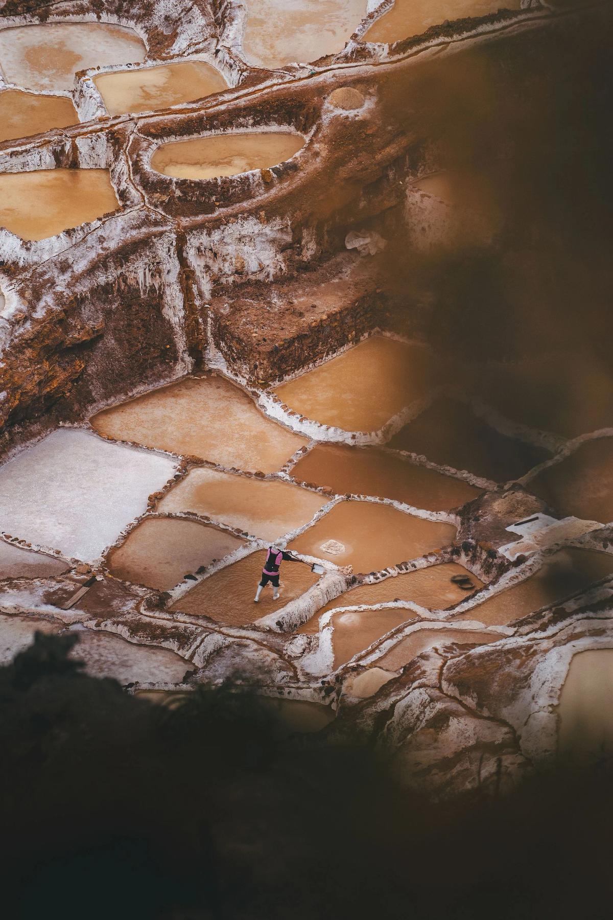 Vista desde arriba, a través de vegetación desenfocada, de un trabajador con botas de jebe rastrillando una poza poco profunda entre las terrazas de las salineras de Maras, en el Valle Sagrado del Perú