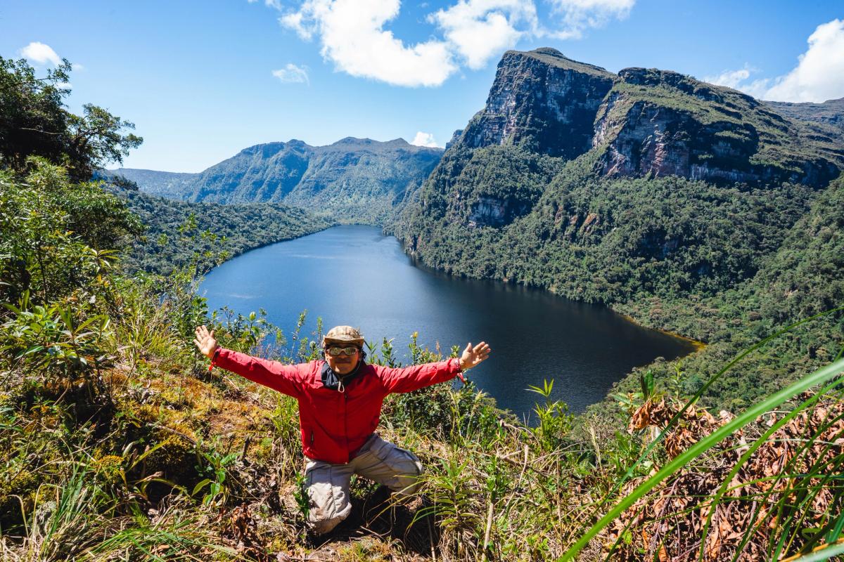 A trekker kneeling with arms outstretched at a viewpoint above Laguna de los Cóndores, with the lake and a tall cliff face behind him on the trail to Quindecocha