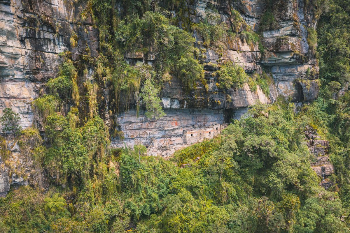 Chachapoya mausoleums built on a narrow ledge beneath a limestone overhang, surrounded by cloud forest above Laguna de los Cóndores