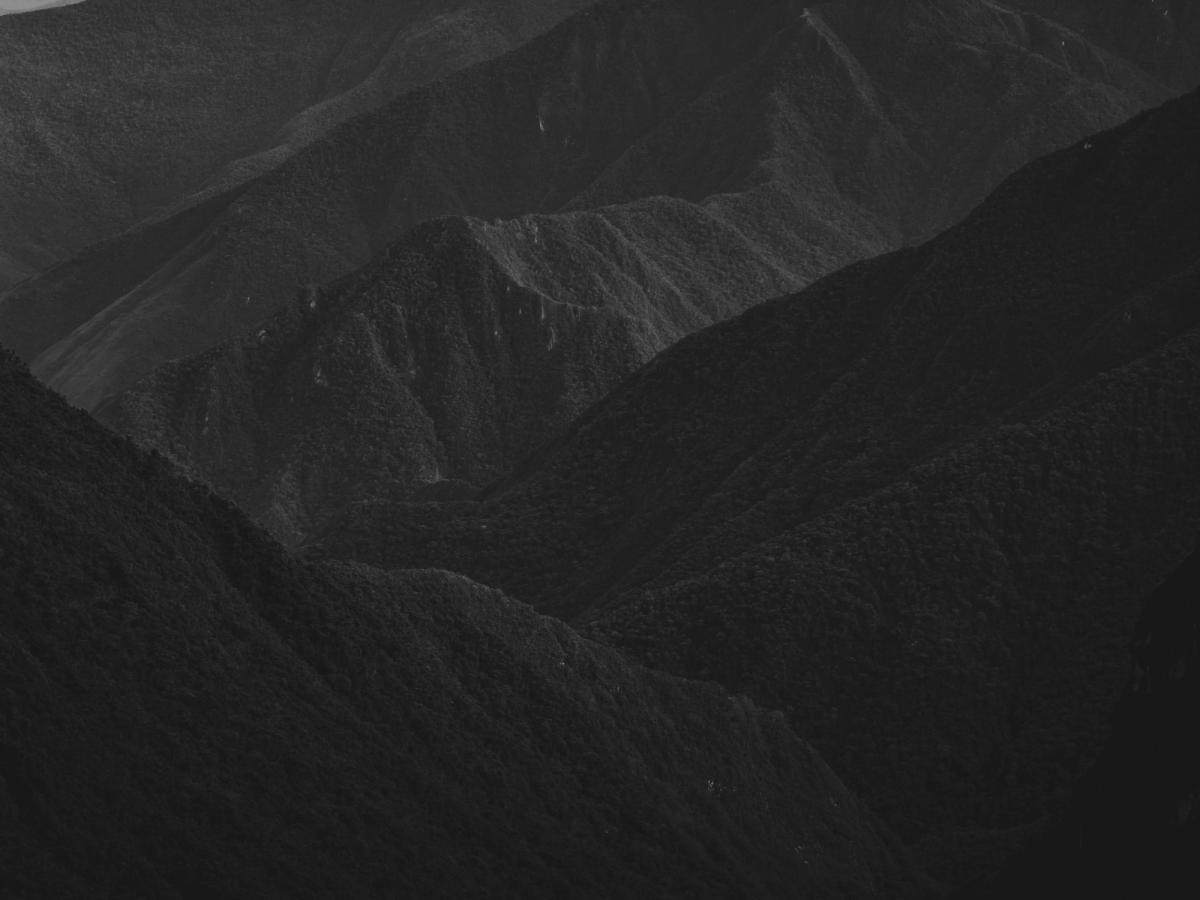 Black-and-white aerial drone view looking down the Ahobamba valley toward Machu Picchu, with layered ridges of dense cloud forest fading into the distance, Cusco region, Peru