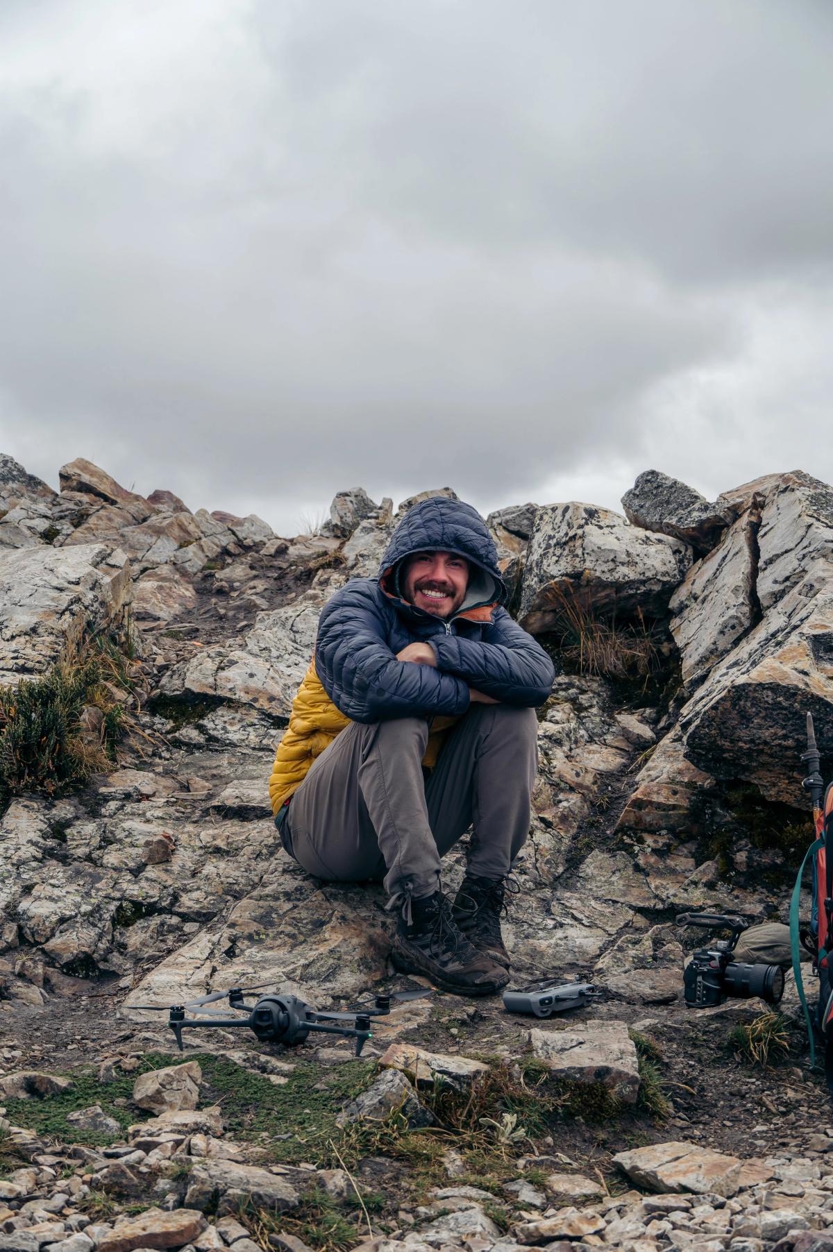 Matt Stacy, fundador de Andes Adventure Visuals, sonríe sentado en una cresta rocosa con una casaca de plumas con capucha bajo un cielo nublado en la Cordillera Huayhuash, con un dron y una cámara en el suelo a su lado