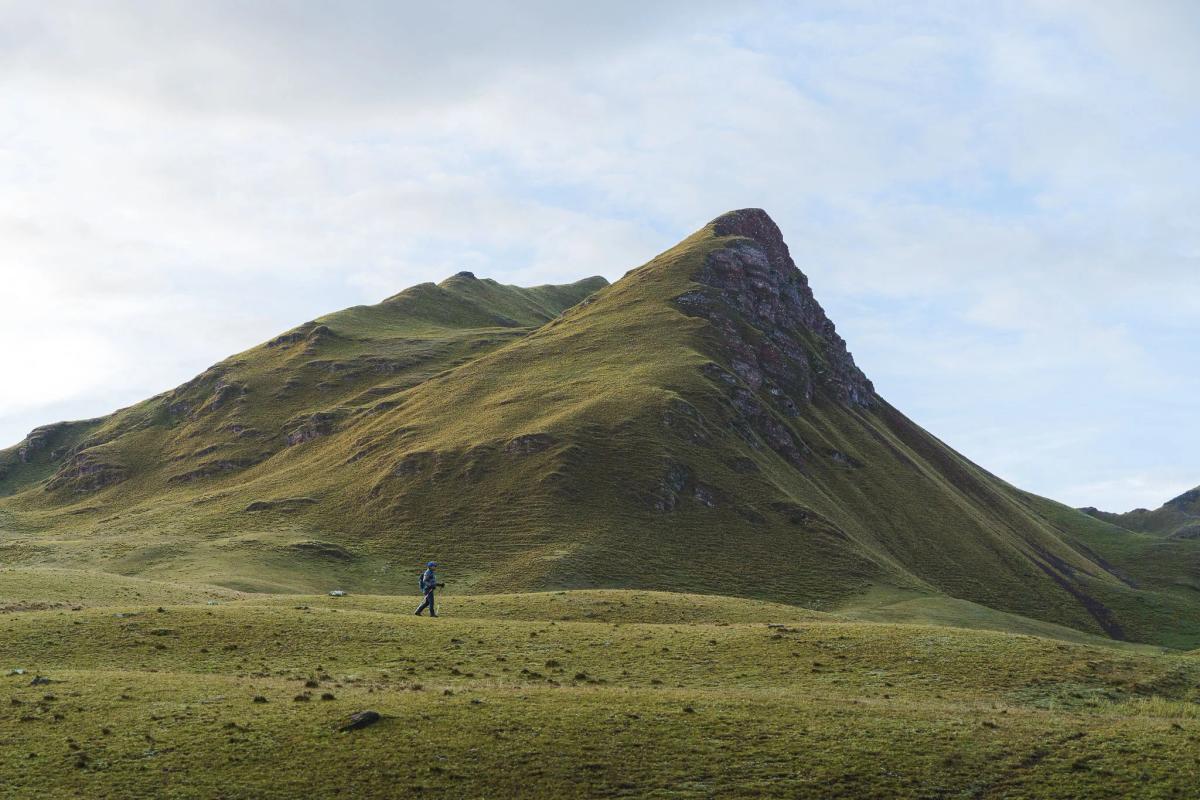 Un excursionista solitario con bastones camina por un pastizal verde y ondulado bajo un pico empinado de hierba con cumbre rocosa en la expedición a Ancascocha, sobre el Valle Sagrado del Perú