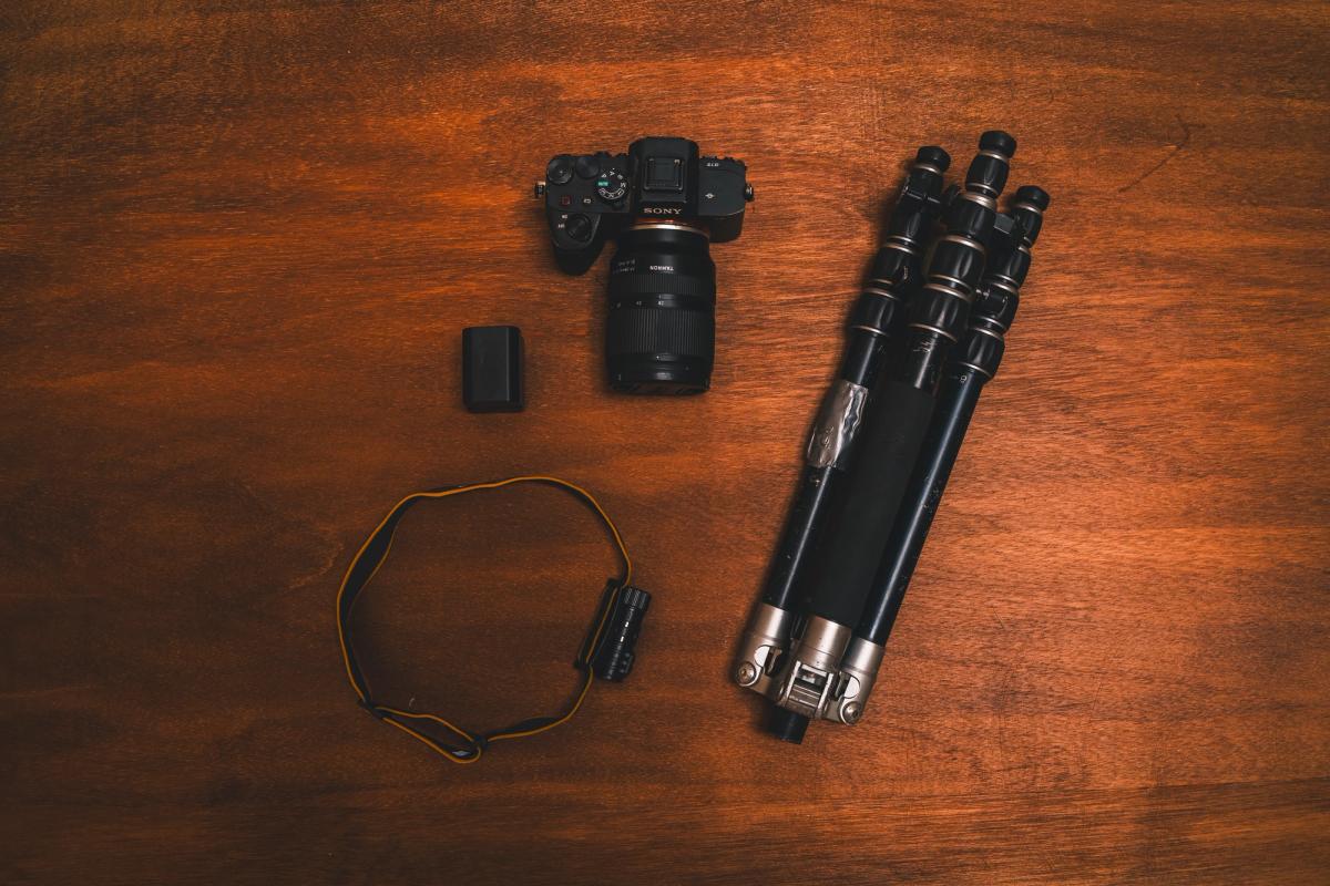 El equipo básico de Matt Stacy para una mañana de taller de fotografía, sobre una mesa de madera: una cámara sin espejo con un lente zoom gran angular, una batería extra, una linterna frontal y un trípode bien usado con cinta adhesiva