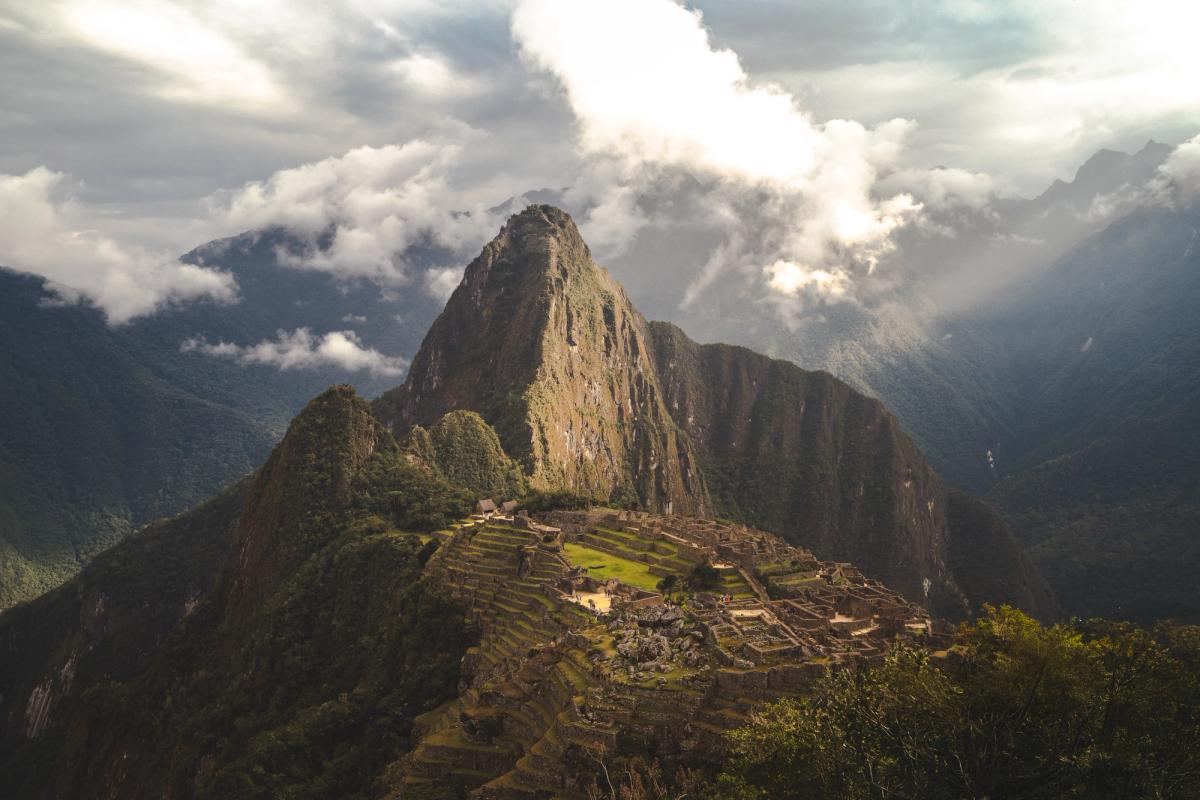 Andenes y construcciones de piedra de Machu Picchu iluminados por un claro entre las nubes, con el Huayna Picchu detrás y montañas de bosque nuboso alrededor, al final del trek Ahobamba, Perú