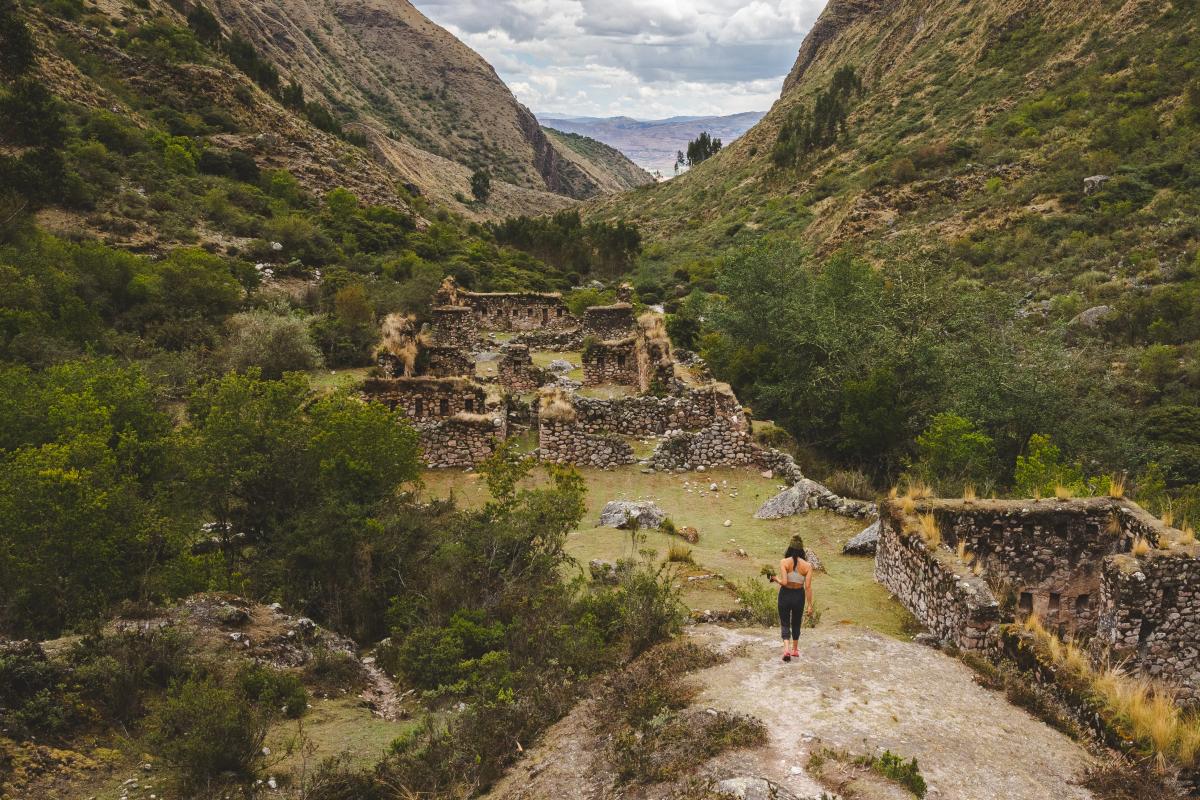 Una excursionista camina por un afloramiento rocoso hacia unas ruinas incas de piedra cubiertas de vegetación, en el fondo de un valle verde y angosto sobre Urubamba, durante un taller fotográfico de AAV en el Valle Sagrado del Perú