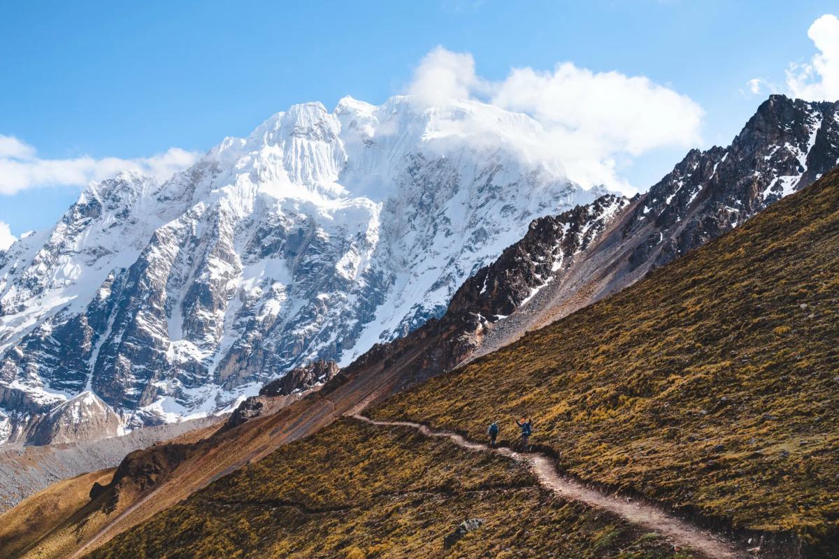 Two AAV trekkers on a narrow trail across a golden grassy slope, one waving, below the massive glaciated face of Nevado Salkantay under a blue sky, Day 1 of the Ahobamba trek, Peru