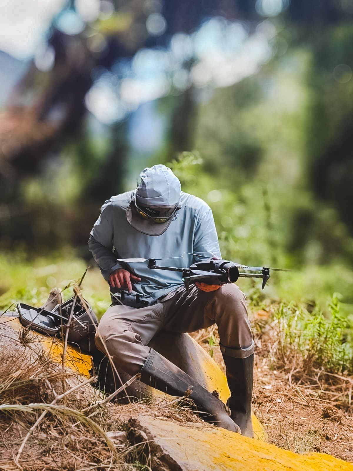 A photographer in a sun hoodie and rubber boots preparing a DJI Mavic drone for launch on a grassy slope above the cloud forest