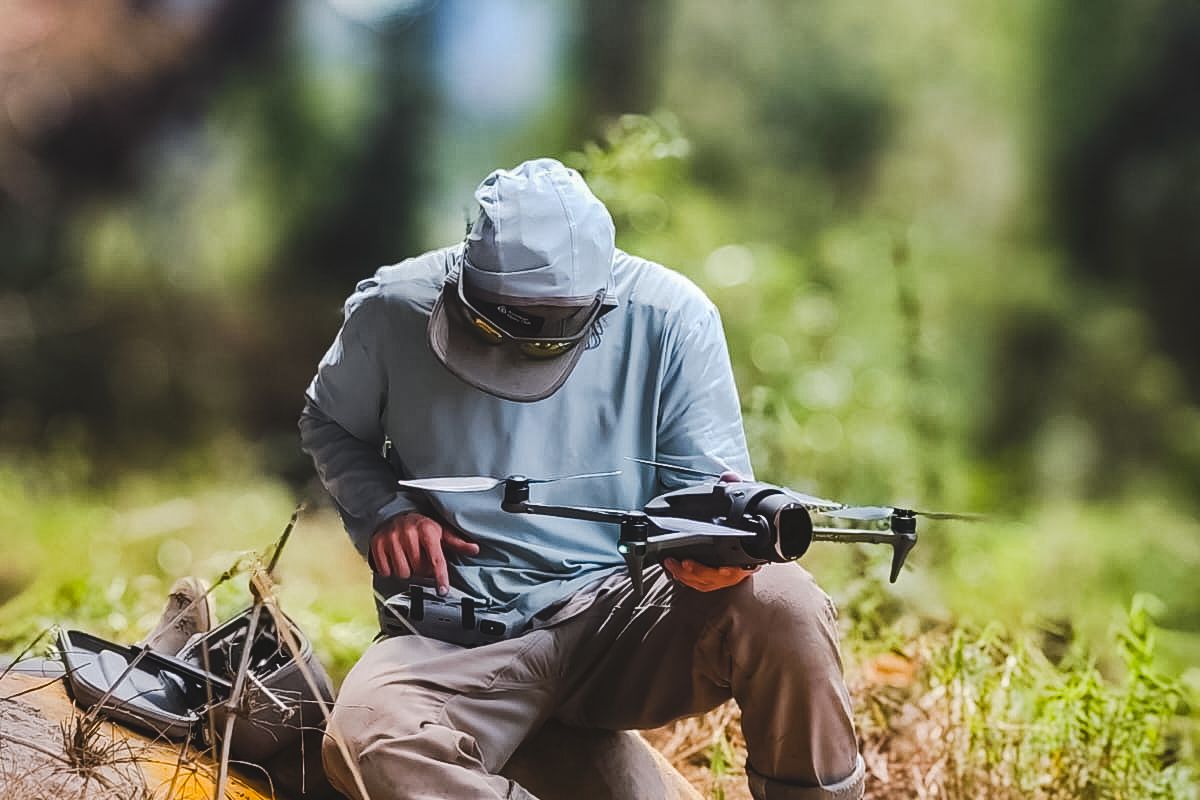 A photographer in a sun hoodie and rubber boots preparing a DJI Mavic drone for launch on a grassy slope above the cloud forest