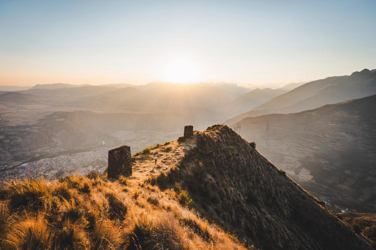 La luz dorada del atardecer ilumina una cresta con pastizales, dos torres incas de piedra llamadas saywas y un excursionista solitario durante un tour fotográfico de AAV, muy por encima de Urubamba y el río en el Valle Sagrado del Perú